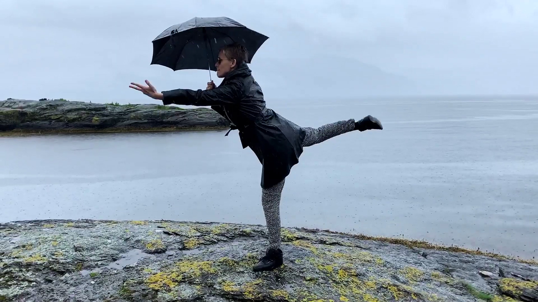 Frau in schwarzer Kleidung und mit Regenschirm tanzt am Meer