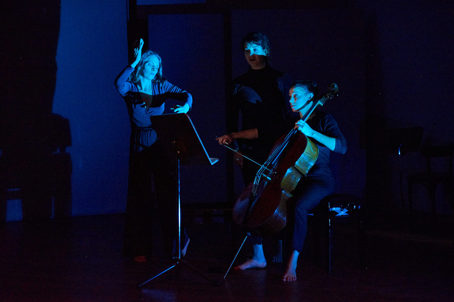 In blauem Licht tanzt und gebärdet Kassandra Wedel, neben ihr steht Maren Schwier und singt. Rechts von ihnen sitzt Larissa Nagel und spielt Cello. SIe alle teilen sich einen Notenständer vor ihnen.