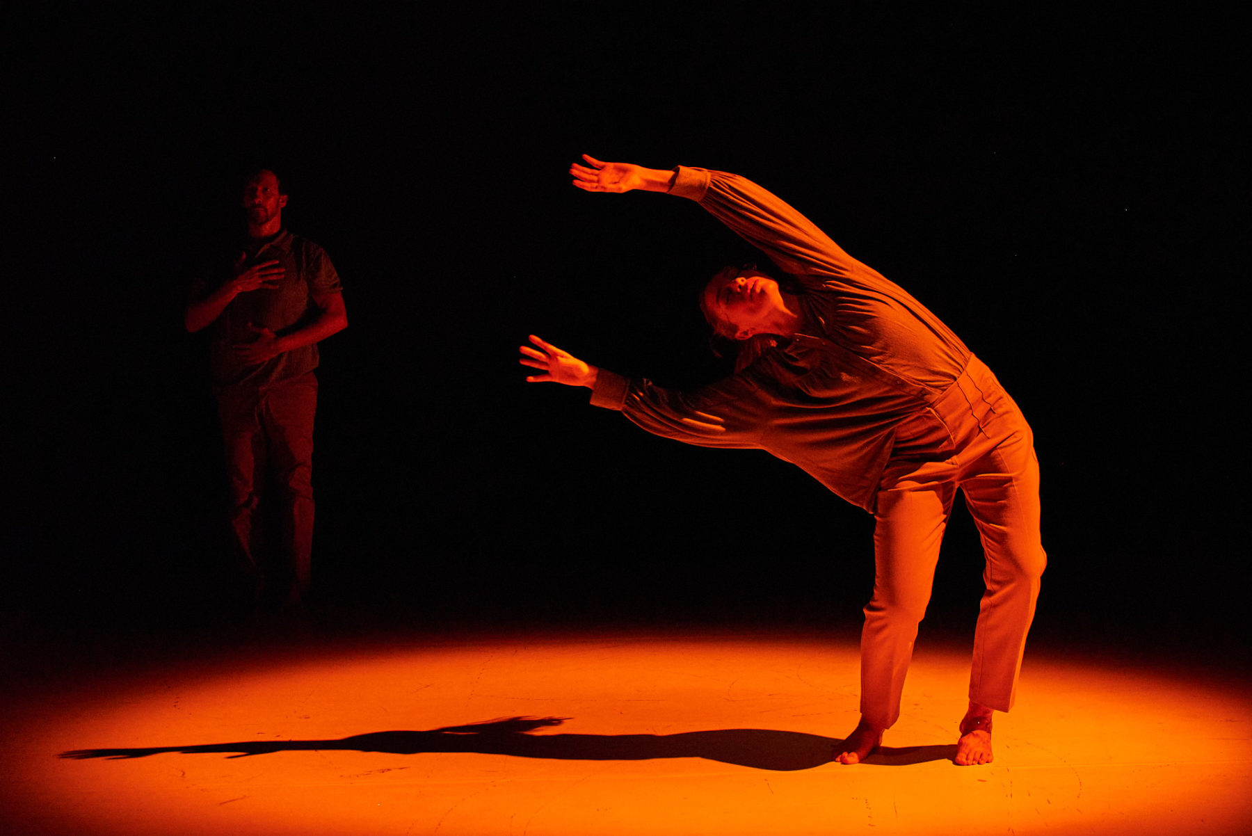 Pilar Andrés tanzt ein Solo in orangenem Licht. SIe steht auf dem Boden und hat ihren Körper nach rechts geneigt, dass der Oberkörper fast horizontal ausgerichtet ist. Im dunklen Hintergrund links hinter ihr steht Mikel Aristegui, welcher eine Hand auf seine Brust und eine auf seinen Bauch hält. Er schaut nach vorne.
