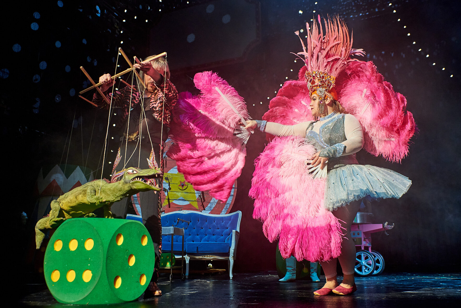 Eine Theaterbühne. Rechts steht eine Person im hellblauen Tutu, die einen Fächer aus großen, pinkfarbenen Federn in der Hand hält. Links liegt ein großer, grüner SChaumstoffwürfel, auf dem eine Krokodil-Marionette balanciert. Hinter dem Würfel steht der Puppenspieler.