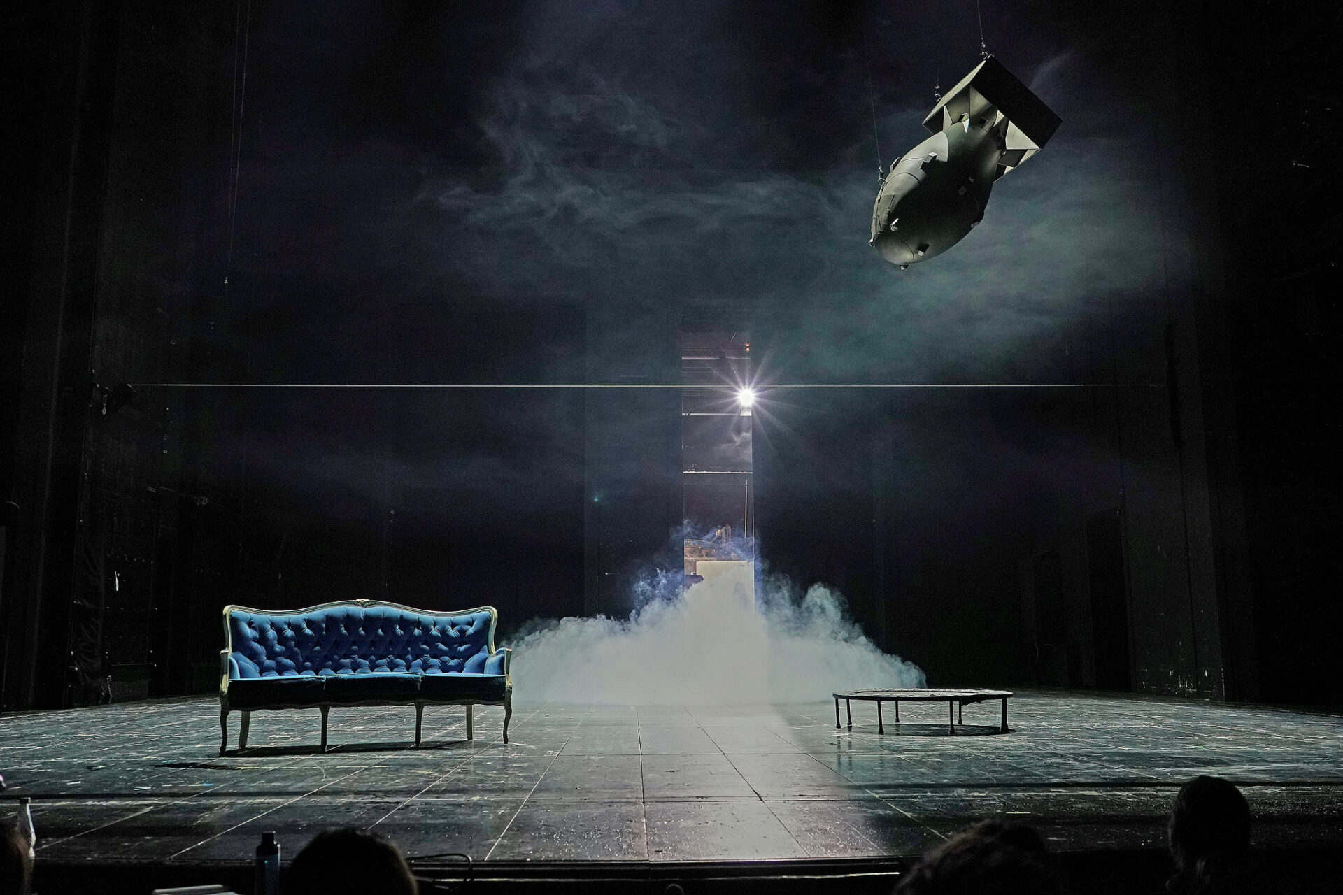 Eine fast leere, schwarz gestrichenen Bühne. Links steht ein blaues Sofa, rechts hängt eine Bombe in der Luft. Im Hintergrund wabert Bühnennebel.