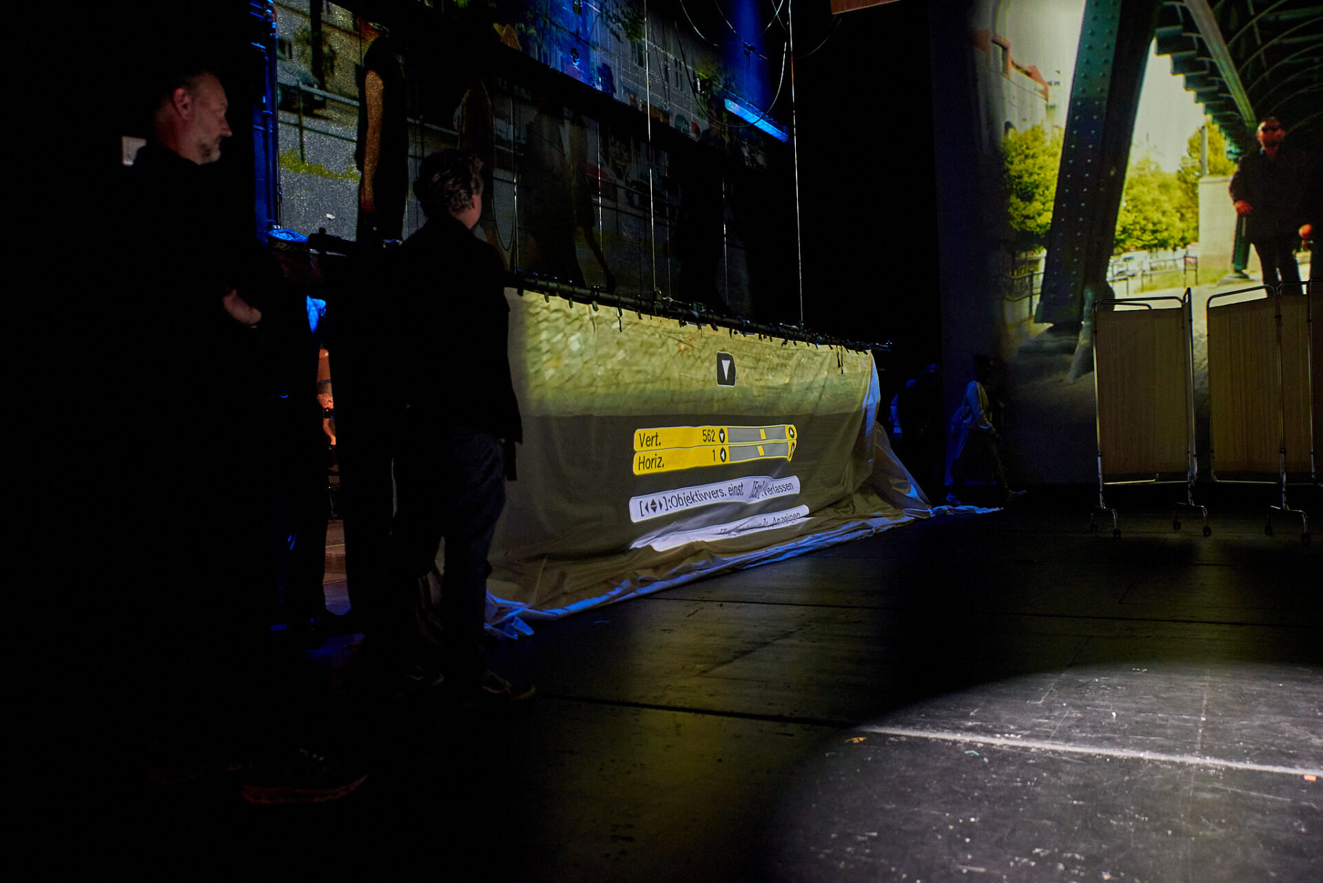 Auf der linken Seite der Bühne stehen zwei Bühnentechniker und beobachten prüfend den Hänger mit der gelblichen Projektionsleinwand, die parallel zur linken Bühnenseite ausgerichtet ist. Im Hintergrund zeichnet sich die linke Hälfte der mittleren Leinwand ab, auf welche eine Filmsequenzen großformatig projiziert wird. 