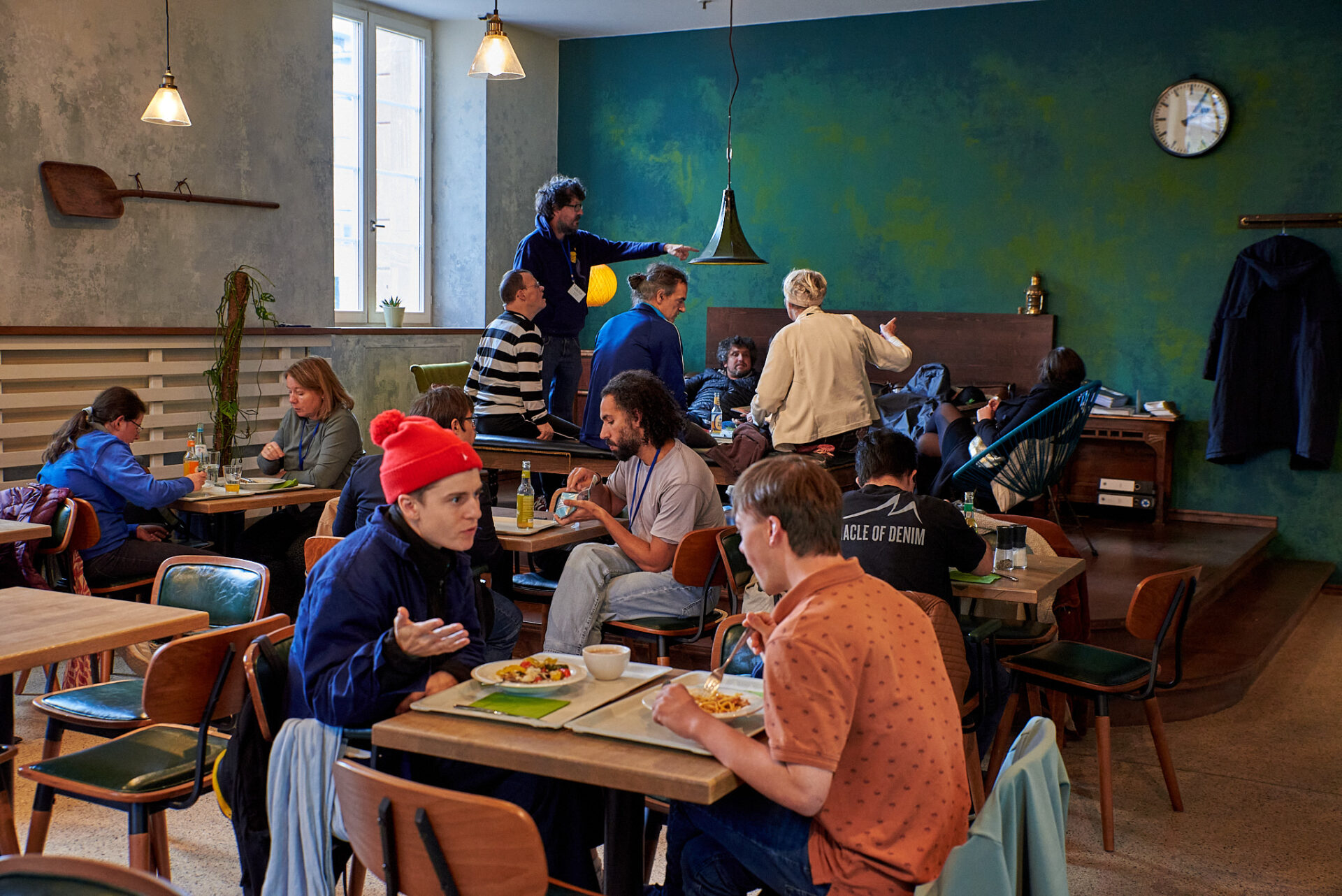 Ein Raum mit einer grün-blauen Wand, warmen Deckenlicht, Tisch- und Stuhlgruppen, an denen Menschen sitzen, essen und sich unterhalten.