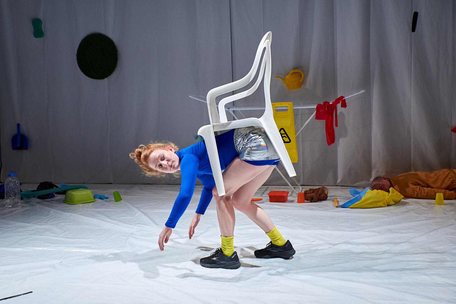 eine Tänzerin mit rotem Haar, blauen Oberteil und silbernen Shorts balanciert einen weißen Gartenstuhl auf ihrem Rücken; sie ist gebeugt und allein auf der weißen Bühne; im Hintergrund einige angehäufte Dinge