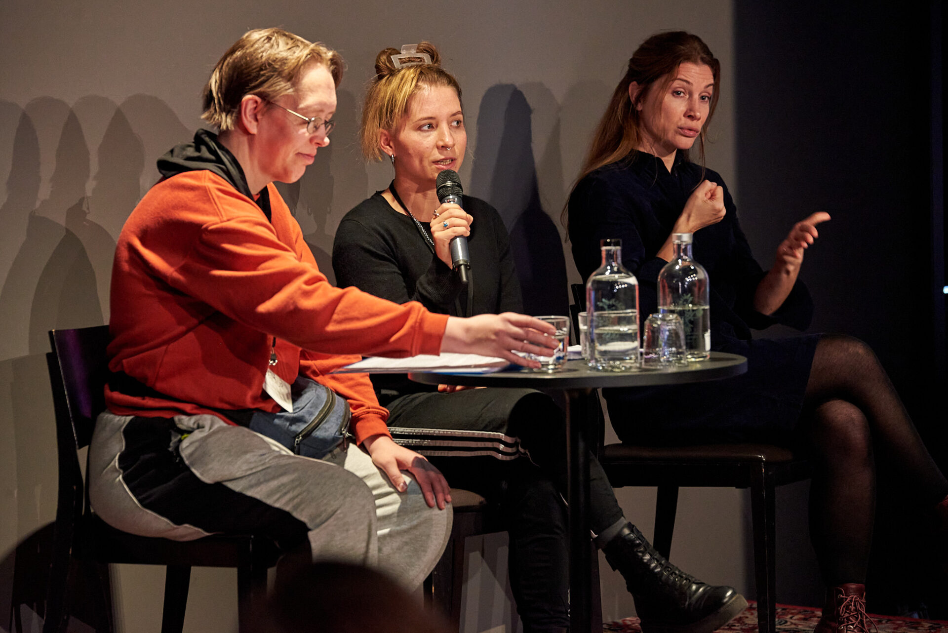 Friederike Jaglitz, Simone Burckhardt und Sarah Verena Bockers sitzen an einem runden Tisch auf dem Podium. Auf dem Tisch stehen zwei Glasflaschen mit Wasser, Friederike Jaglitz greift nach einem Wasserglas.