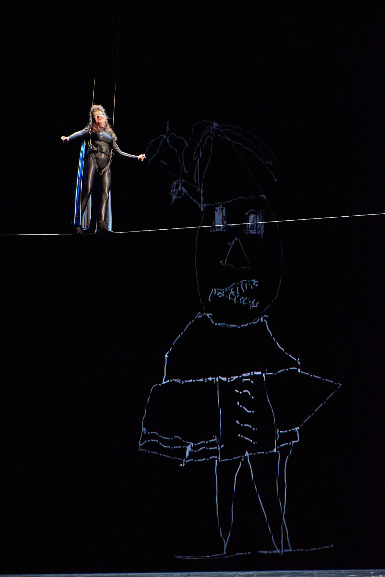 Die Darstellerin steht auf einem Drahtseil und trägt einen Monolog vor. Sie ist mit einem Klettergurt und zwei Seilen gesichert. Sie trägt einen schwarzen Anzug und einen lila Umhang. Es trägt ein Superheldenkostüm mit Umhang. Auf der schwarzen Leinwand im Hintergrund sieht man eine gezeichnete Figur, die auf das Bild "Angelus Novus" von Paul Klee verweist