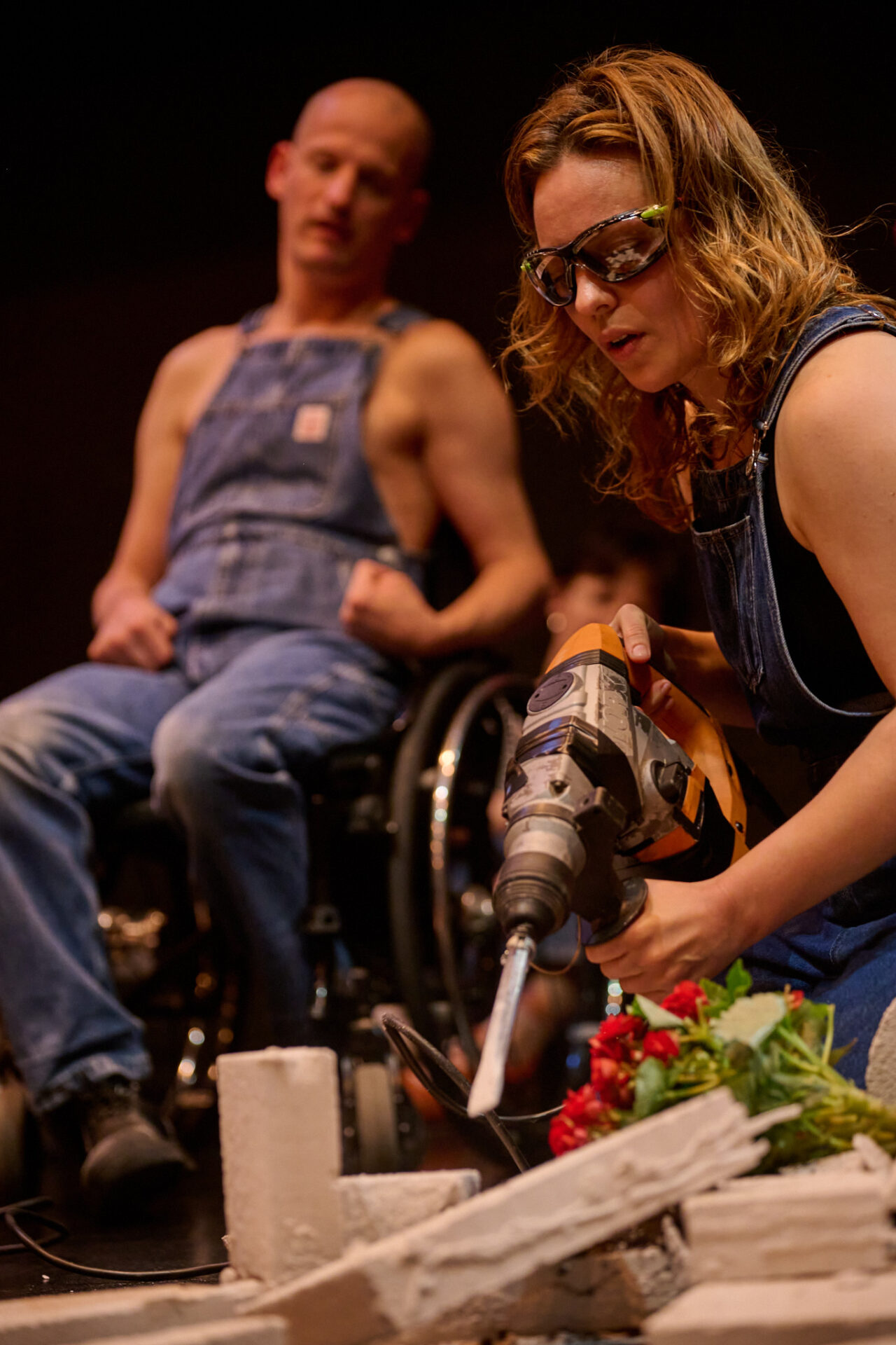 Eine Performerin in blauer Latzhose und Schutzbrille bearbeitet mit einem Elektromeißel einen weißen Steinquader, aus dem rote und weiße Blumen hervortreten. Im Hintergrund sitzt Michael Turinsky in einer blauen Latzhose im Rollstuhl und beobachtet die Szene.