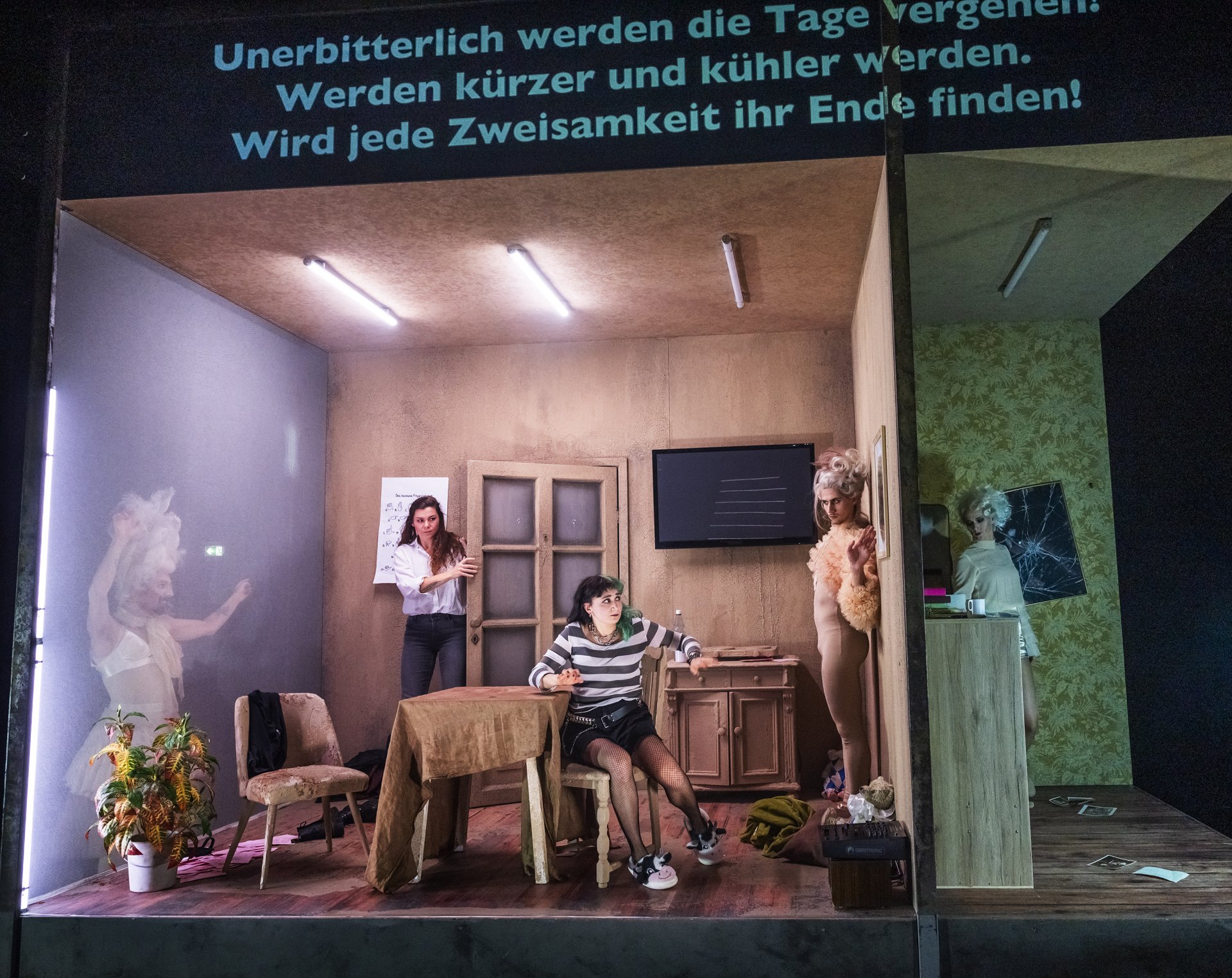 Das Bild zeigt das Bühnenbild von "Altbau in zentraler Lage". Zu sehen sind zwei voneinander abgetrennte Zimmer. Im Zentrum ist ein Esszimmer zu sehen, an dessen Rändern drei Gespenster in enganliegenden Rüschenkostümen lauern. Die Bewohnerin Zoey sitzt verschreckt am Tisch, während ihre Nachbarin Trisha zur Tür reinkommt.